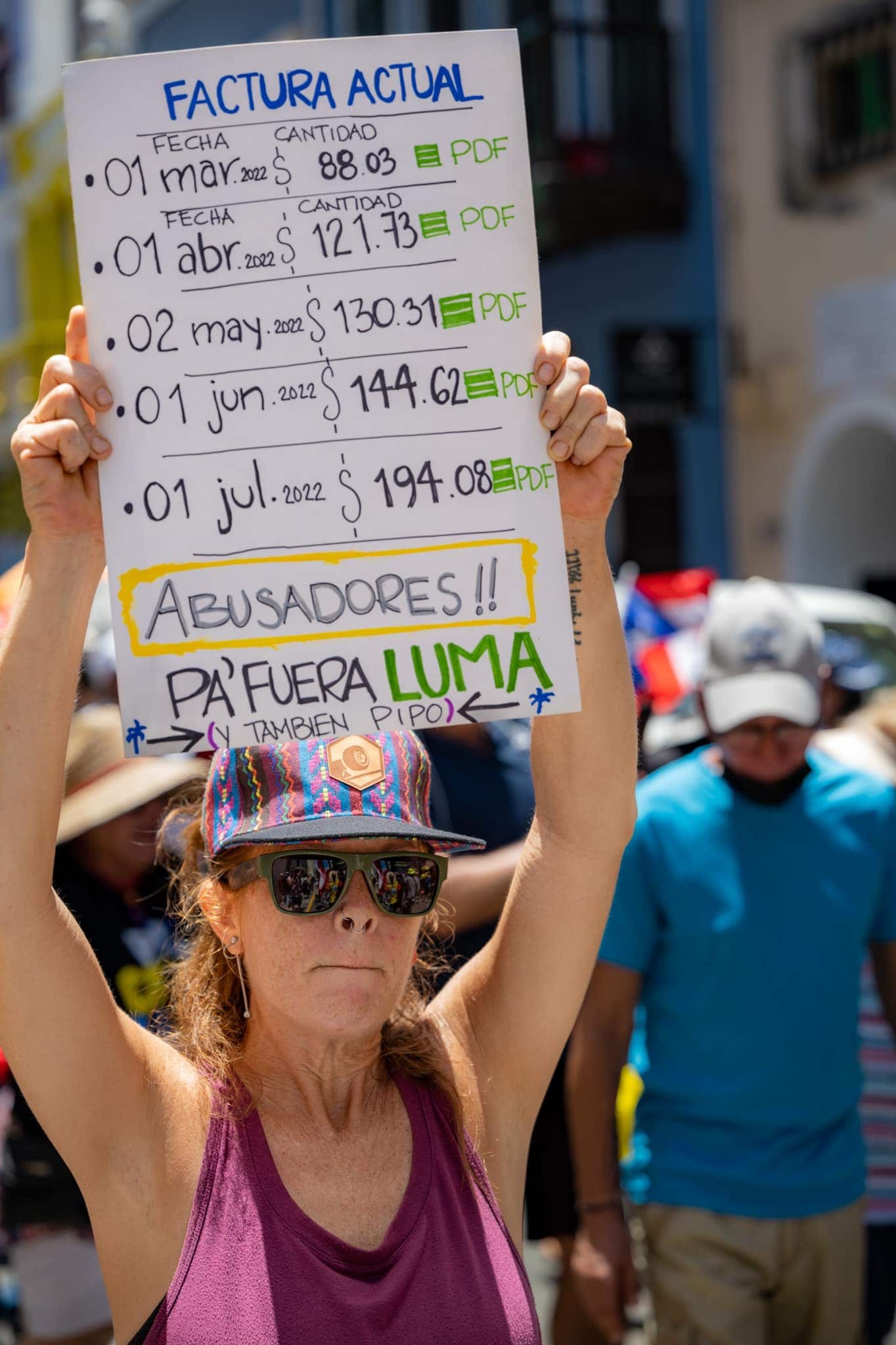 Woman holding a protest sign showing increased electricity bills from March to July 2022, with messages against LUMA Energy in Puerto Rico.