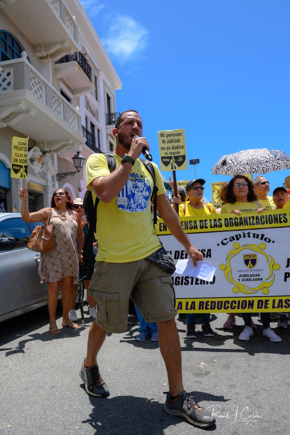 Activist and Vice-president of FMPR with a microphone leading a protest with members of the Federación de Maestros de Puerto Rico, holding signs calling for protection of pensions and against privatization.