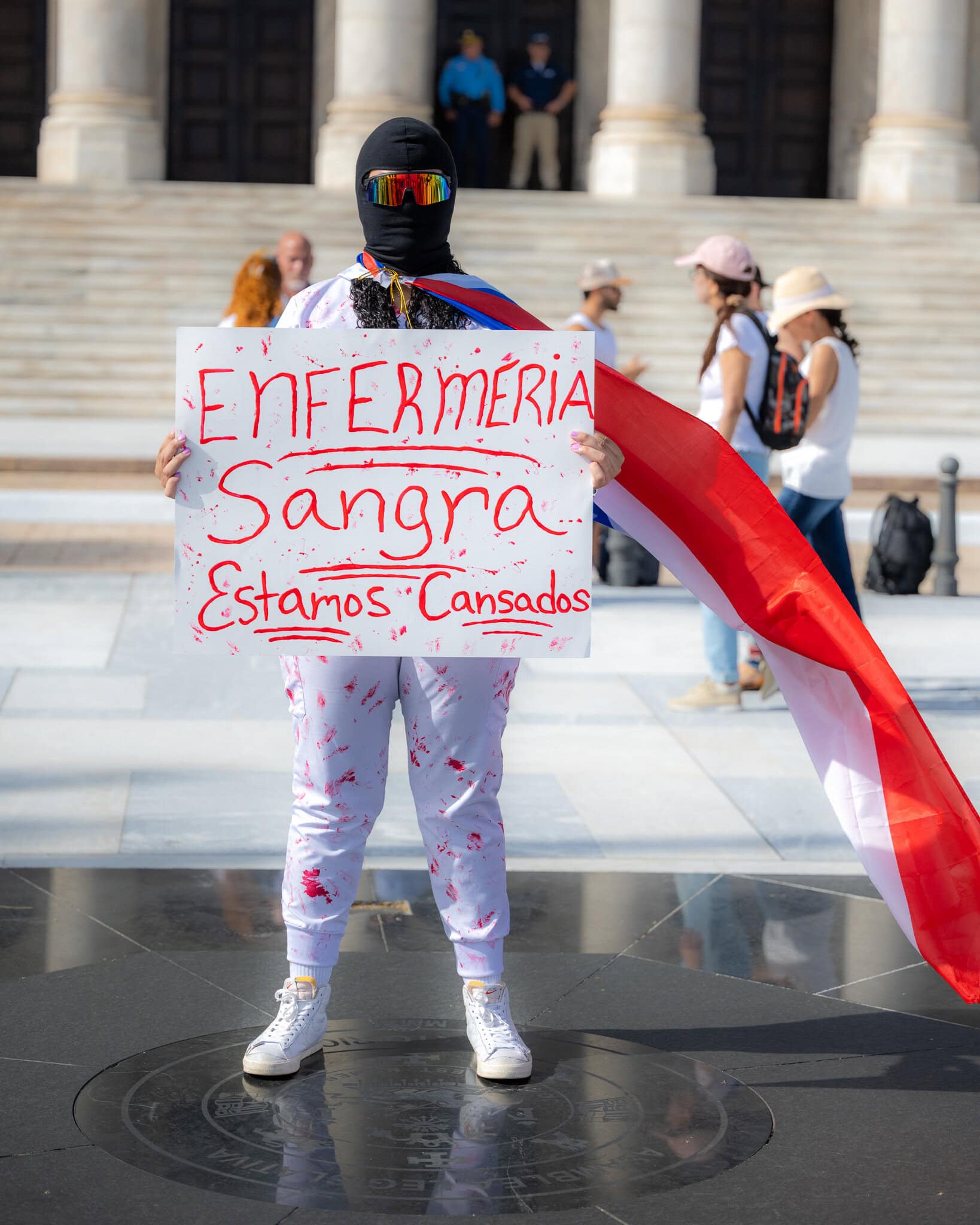 Protester dressed in white holding a sign that reads “Enfermería sangra, estamos cansados” at El Capitolio.