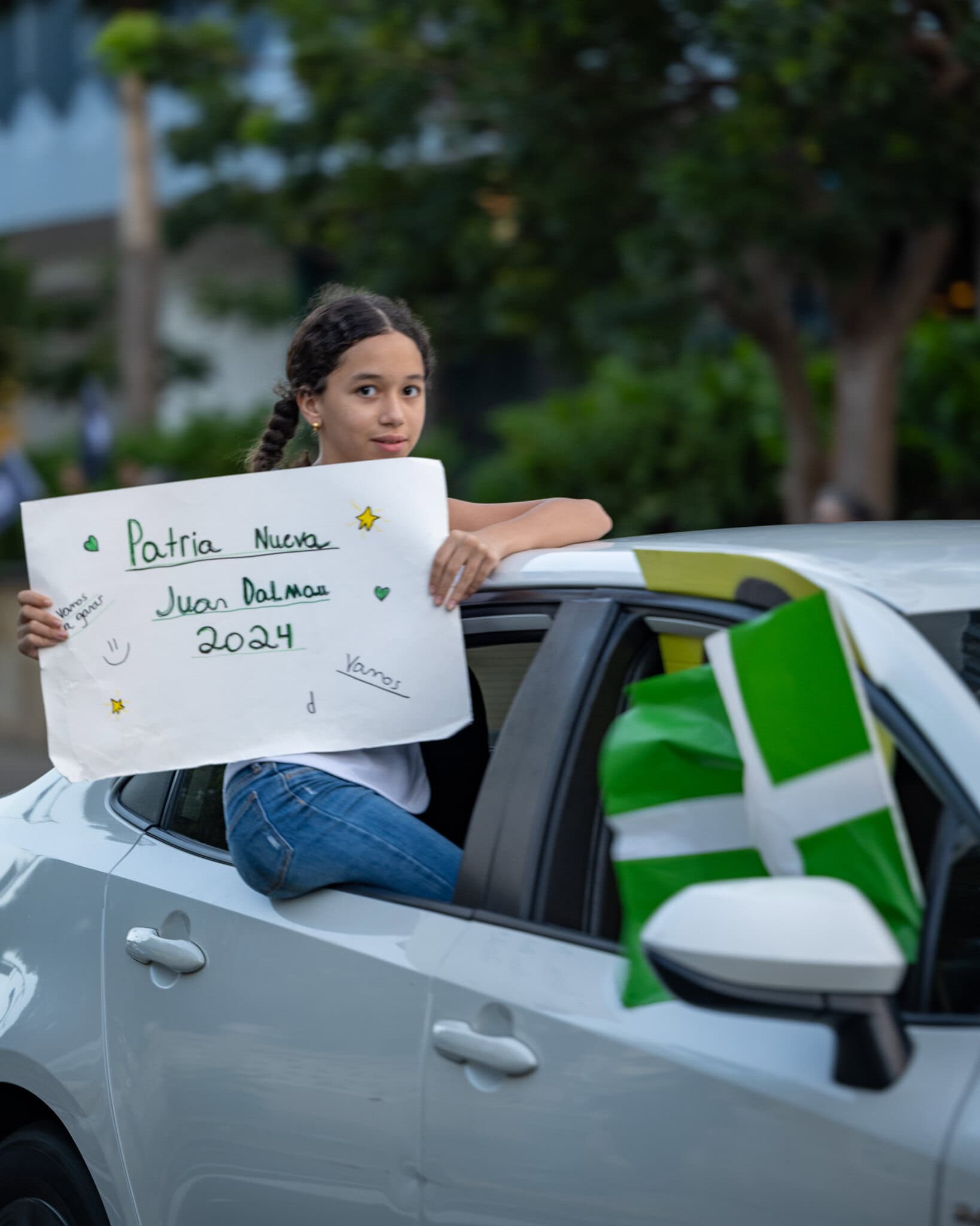 Young girl holding a sign that says 'Patria Nueva Juan Dalmau 2024' out of a car window with a green and white flag.